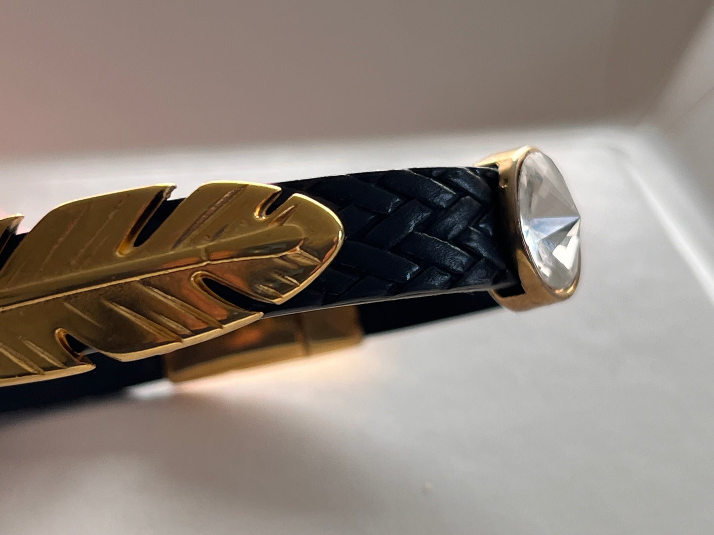 Sort flettet skinnarmbånd med gulldetaljer. Gullbelagt fjær
